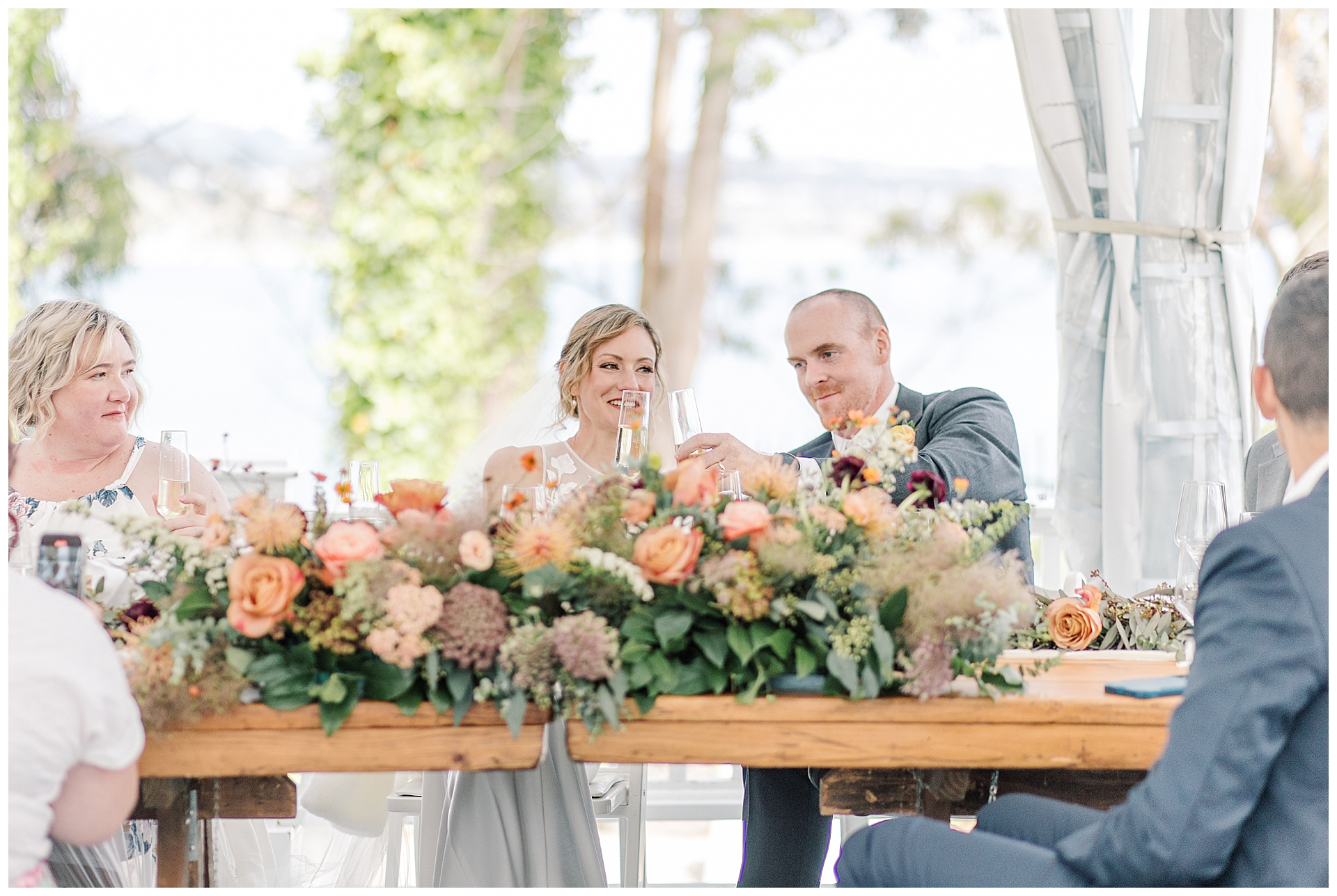 The Bride and groom clink a glass at their head table decorated with orange florals. 